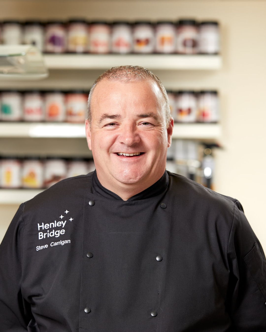 Man wearing a black chef's coat with 'Henley Bridge' branding in a kitchen setting.