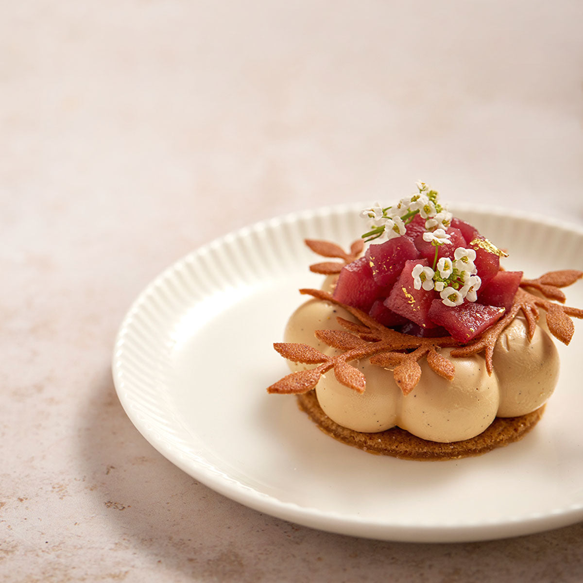 A beautifully plated dessert featuring beige meringue-like dollops topped with pink-red fruit, small white flowers, and brown leaf garnish on a white plate against a light beige background.