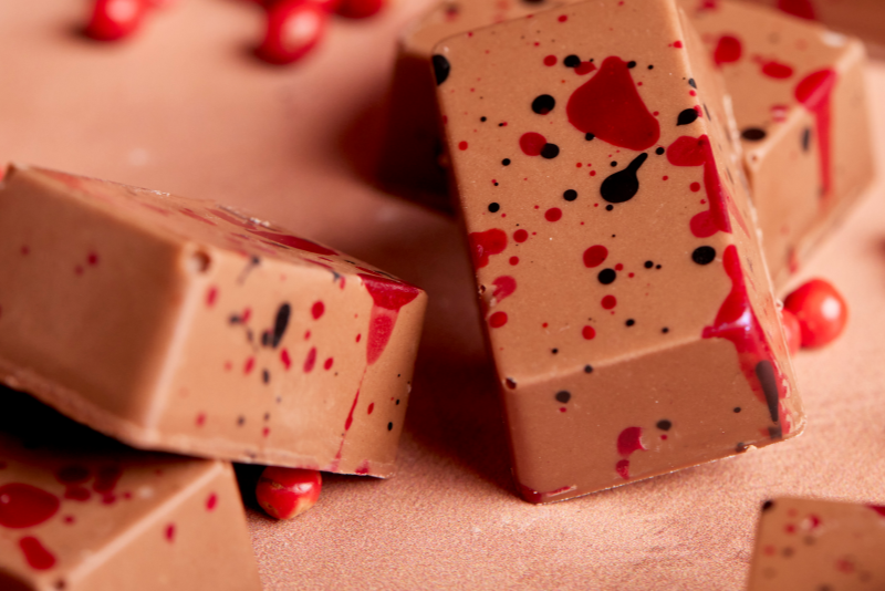 Chocolate-covered rectangular bars with red and black paint splatter details, arranged on a pinkish surface with scattered red berries.